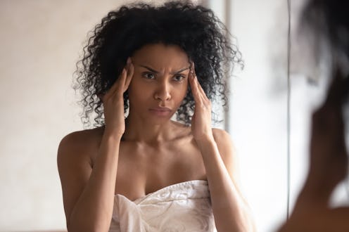 Thoughtful african American woman look in mirror in bathroom worry about facial wrinkles think of an...