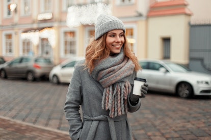 Happy young woman smiling in a trendy coat with a knitted vintage hat and fashionable warm scarf wit...