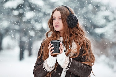 Happy young woman with cup of coffee in snowy winter day. Girl enjoys winter, frosty day. Walk in wi...
