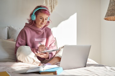 Hipster teen girl school student with pink hair wear headphone write notes watch video online webina...