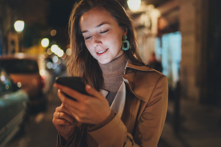 Attractive happy young woman reading good news on her mobile phone during night walk in the city str...