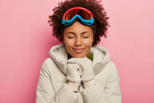Feeling reaxed and happy. Healthy adorable woman with curly hair, wears white down padded coat and m...