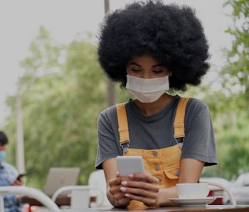 African American young hipster gen z female with Afro hair wearing face mask holding smart phone usi...