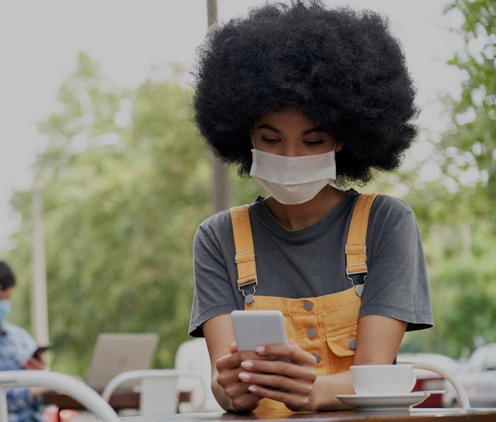 African American young hipster gen z female with Afro hair wearing face mask holding smart phone usi...
