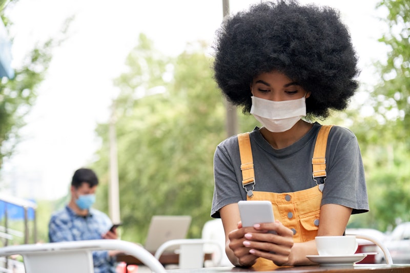 African American young hipster gen z female with Afro hair wearing face mask holding smart phone usi...