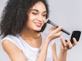 Smiling afro american woman applying makeup powder or foundation with brush isolated on a grey backg...