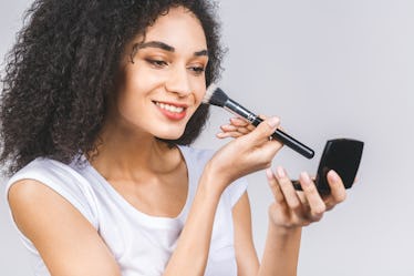 Smiling afro american woman applying makeup powder or foundation with brush isolated on a grey backg...