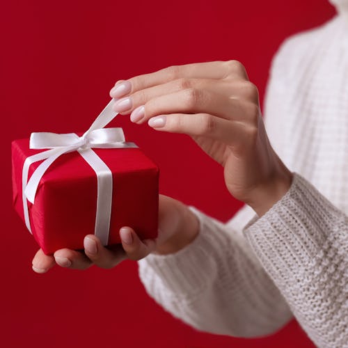 Female hands open Christmas red gift box with white bow on the red background. White sweater. New Ye...