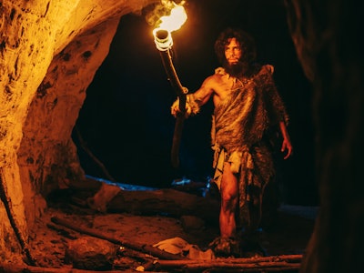 Primeval Caveman Wearing Animal Skin Exploring Cave At Night, Holding Torch with Fire Looking at Dra...