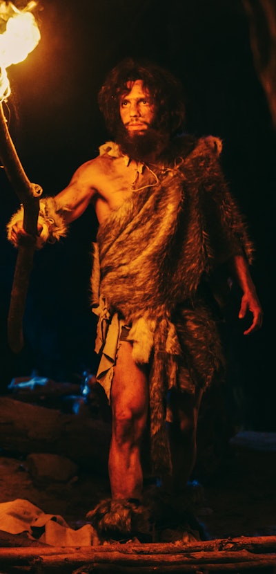 Primeval Caveman Wearing Animal Skin Exploring Cave At Night, Holding Torch with Fire Looking at Dra...