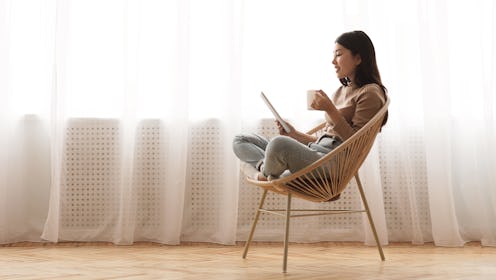 Free time. Girl using tablet and drinking coffee, sitting in wicker chair against window, panorama w...