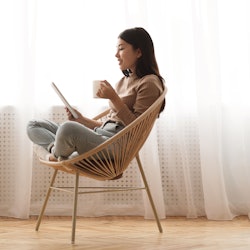 Free time. Girl using tablet and drinking coffee, sitting in wicker chair against window, panorama w...