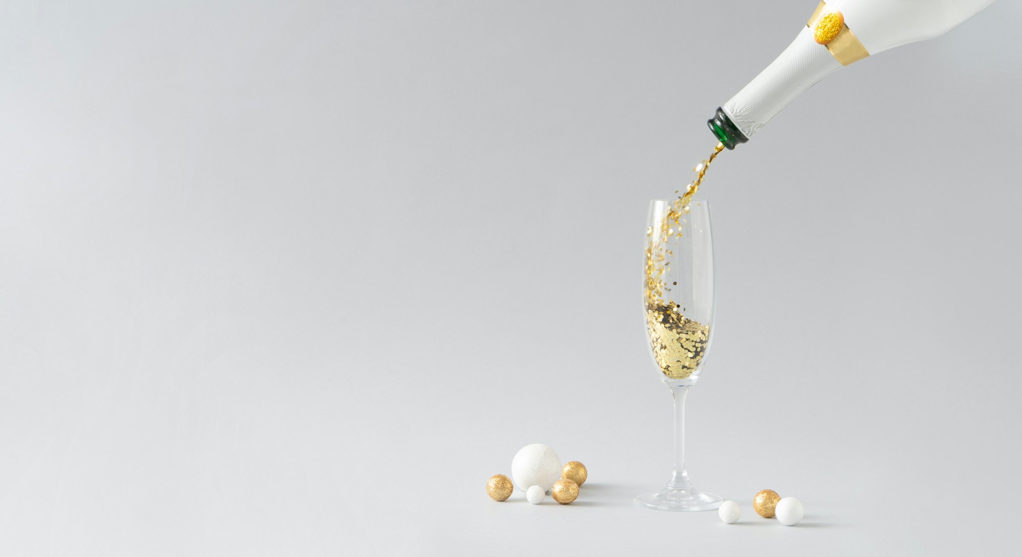 Champagne bottle pouring golden glitter into the glass with white and golden decoration against grey...