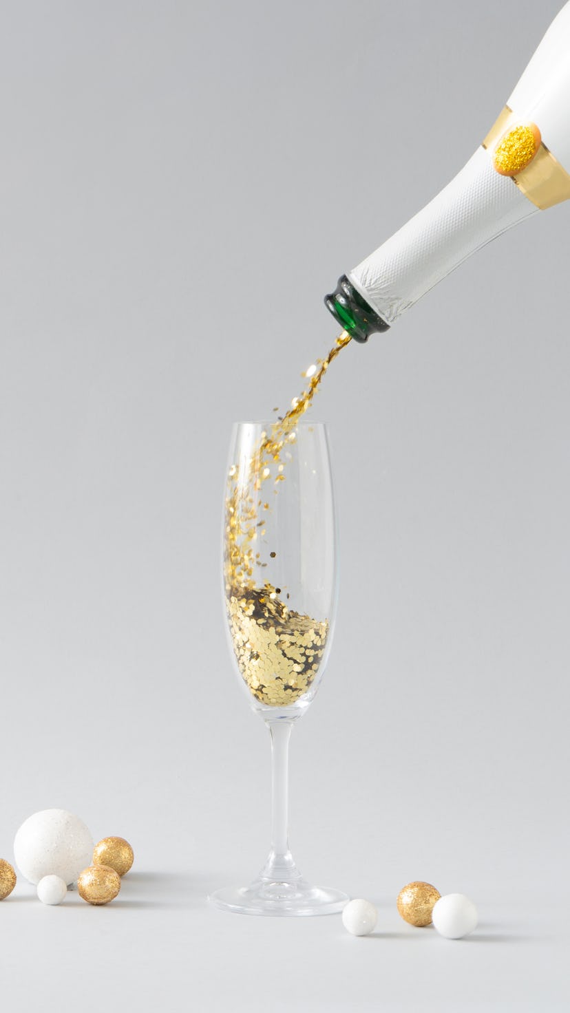 Champagne bottle pouring golden glitter into the glass with white and golden decoration against grey...