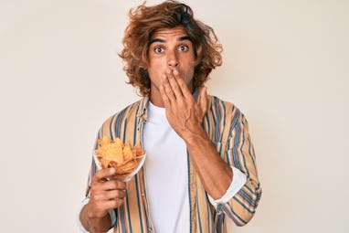 Young hispanic man holding nachos potato chips covering mouth with hand, shocked and afraid for mist...