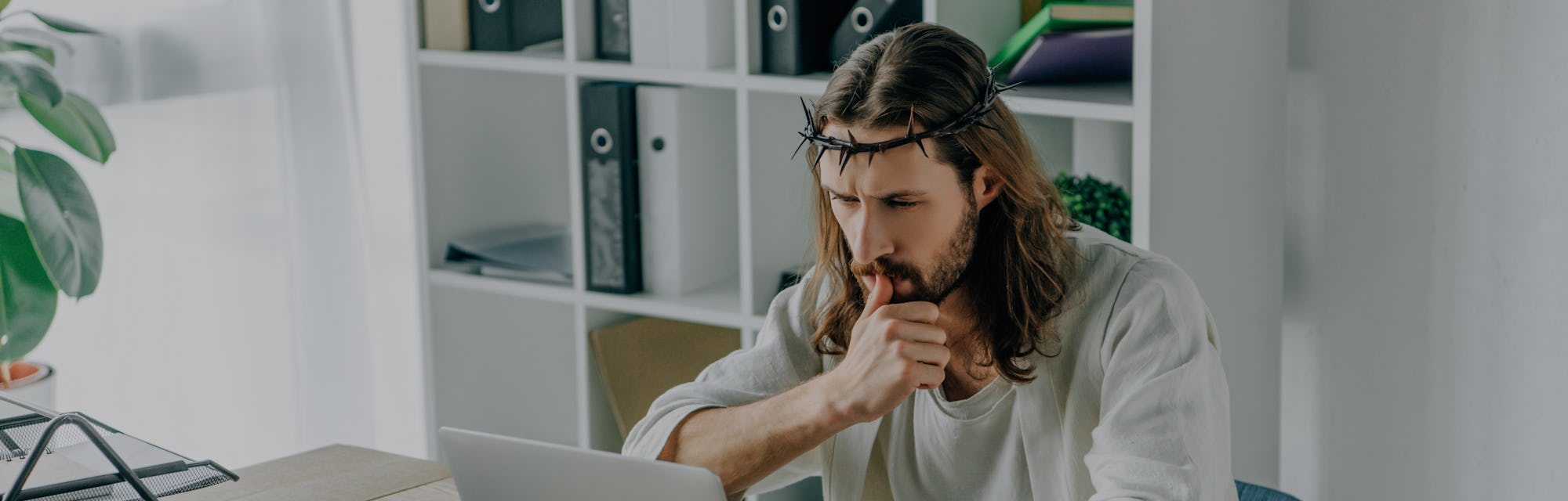 pensive Jesus in crown of thorns using laptop at table in modern office