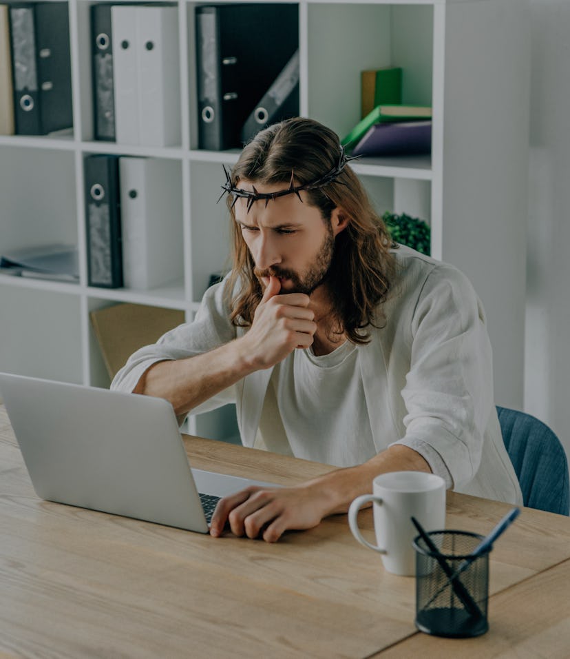 pensive Jesus in crown of thorns using laptop at table in modern office
