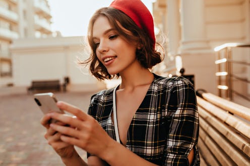 Winsome elegant girl texting message in autumn day. Debonair short-haired woman in red beret standin...