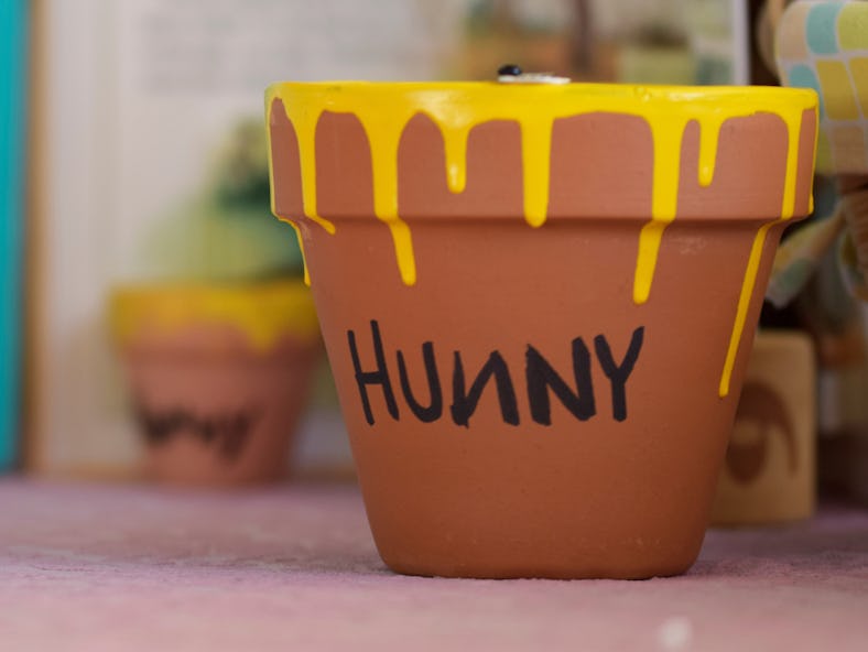 A small clay pot dripped with yellow paint and "hunny" written, with a children's book blurred behin...