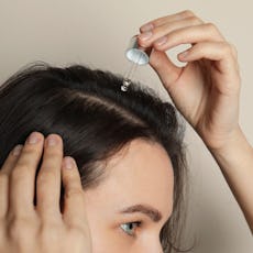 Woman applying oil onto hair on grey background, closeup. Baldness problem