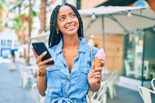 A young woman is outside looking love-struck, with her phone in one hand and an ice cream cone in th...