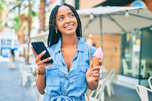 A young woman is outside looking love-struck, with her phone in one hand and an ice cream cone in th...