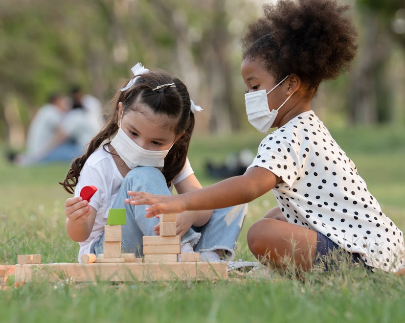 Two young children, each wearing a protective face mask, playing outside together with blocks. 