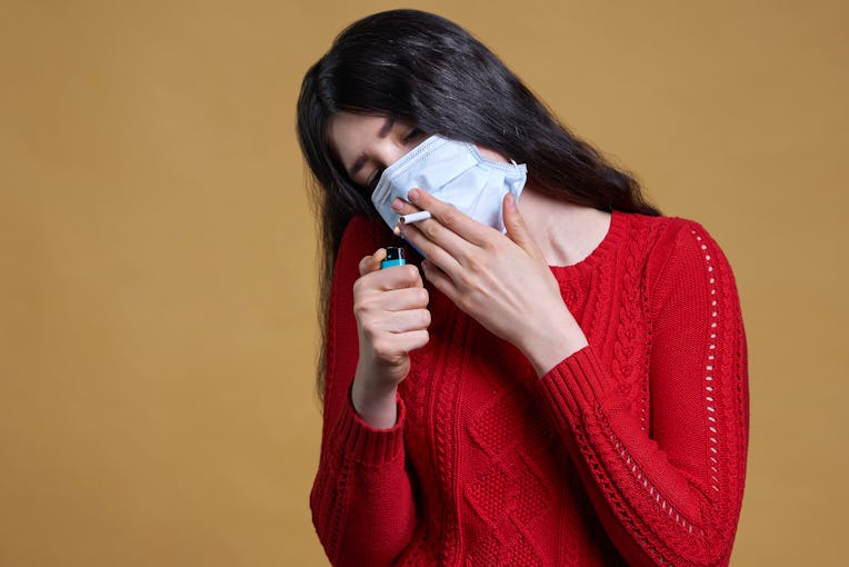 Young girl wearing medical protective mask lights up, holding a cigarette in her mouth through the m...