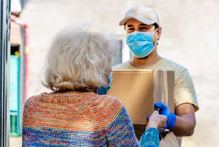 Young male volunteer in mask gives an elderly woman boxes with food near her house. Son man helps a ...