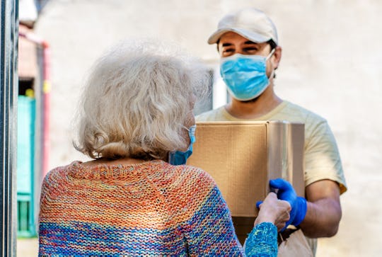 Young male volunteer in mask gives an elderly woman boxes with food near her house. Son man helps a ...