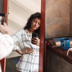 Photo of attractive brunette woman wearing shirt taking selfie photo and looking at mirror in apartm...