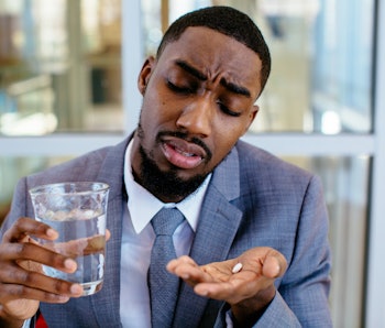 Portrait of a sick young man in business suit taking a medicine pill with water 