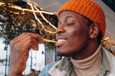 Happy young African-America man in orange knitted hat enjoys eating delicious french fries in contem...