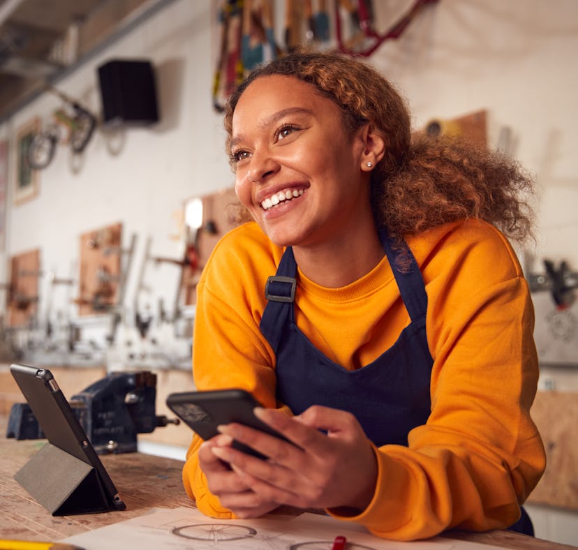 Female Business Owner In Workshop Using Digital Tablet And Holding Mobile Phone