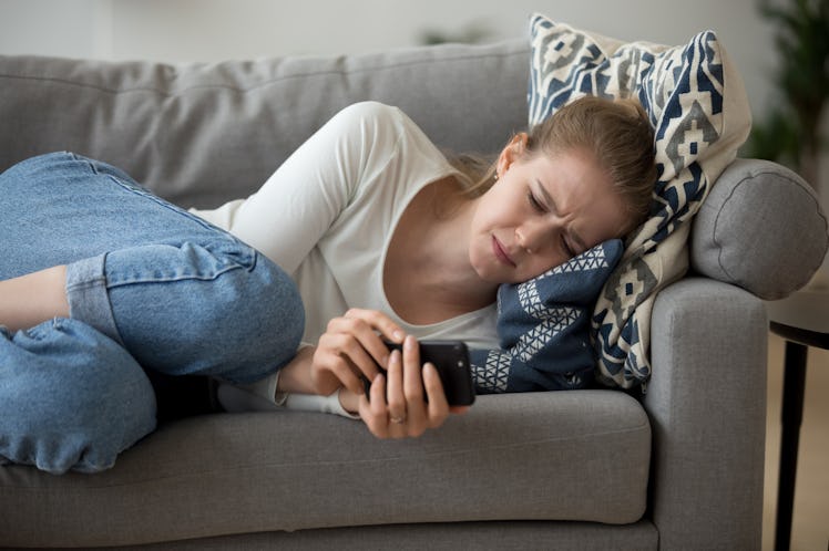 Upset worried young woman crying lying on couch holding phone waiting for call message looking at sm...