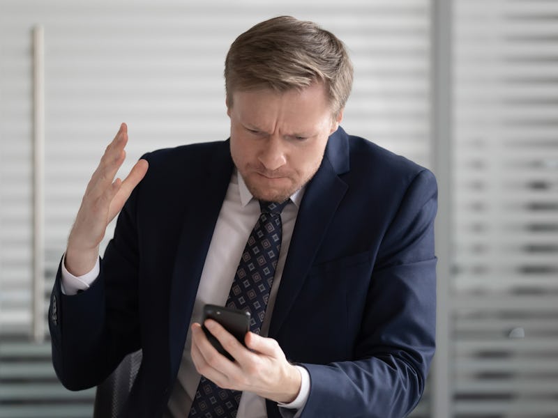 Angry stressed business man wear suit frustrated with broken stuck smartphone in office holding usin...