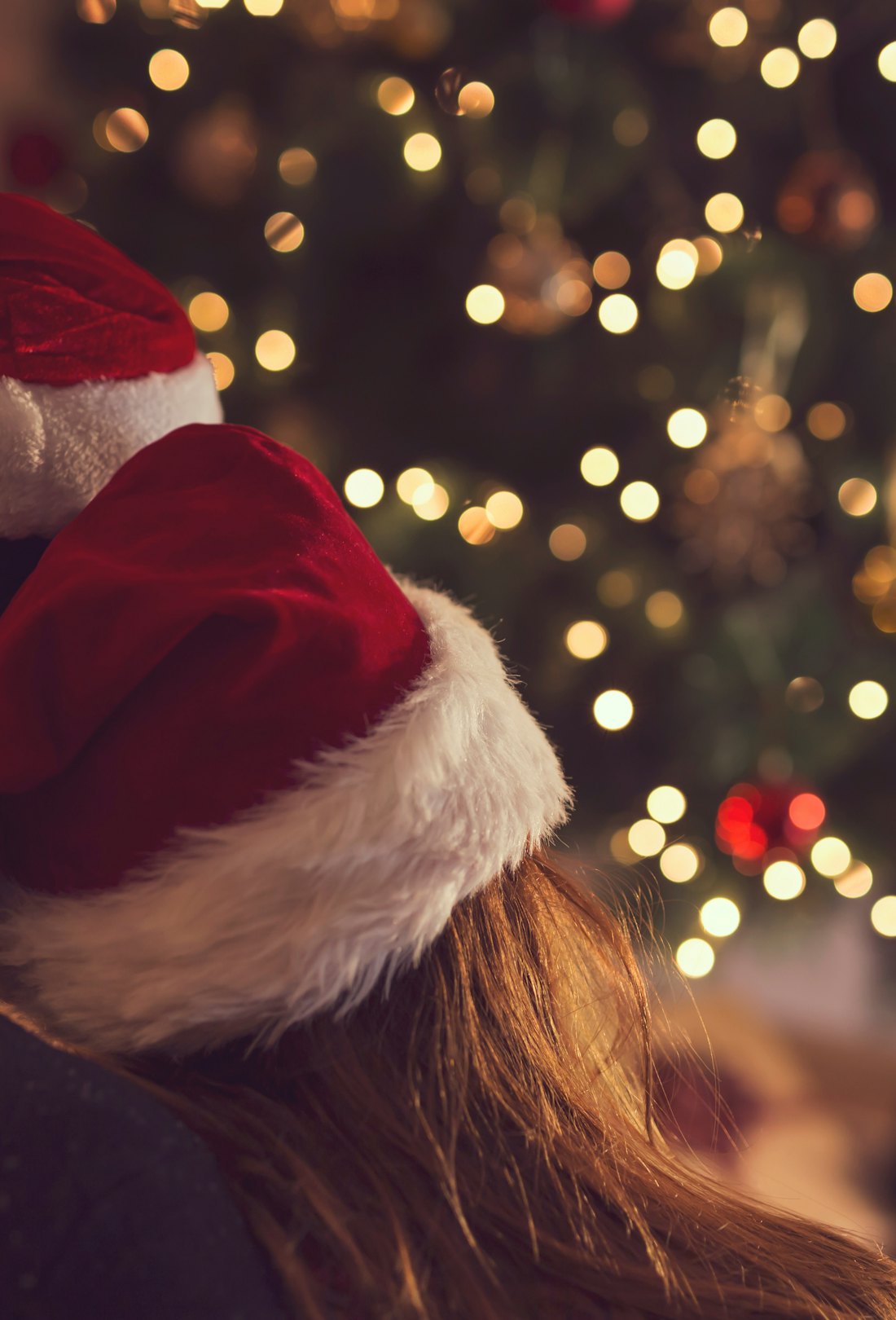Couple in love sitting next to a Christmas tree, wearing Santa's hats, hugging and looking away from...