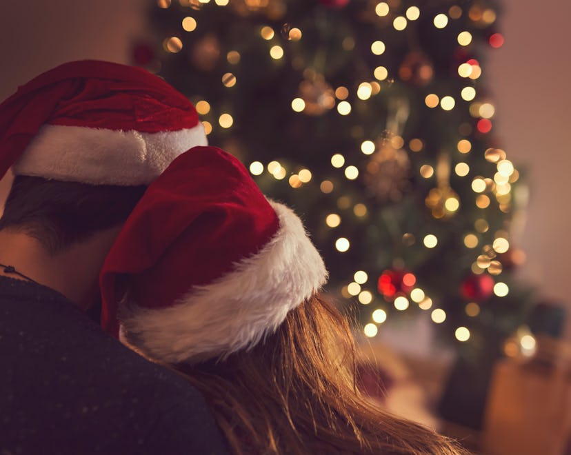 Couple in love sitting next to a Christmas tree, wearing Santa's hats, hugging and looking away from...