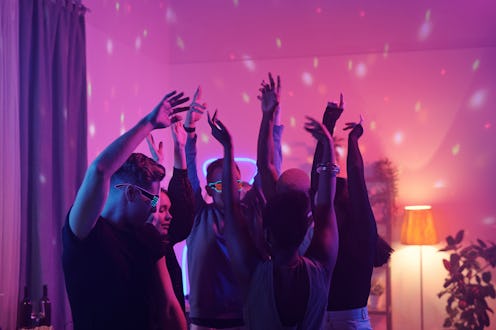 Young intercultural friends in smart casualwear raising arms while dancing together at home party in...