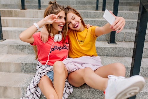 Smiling dark-haired girl making selfie while embracing with stunning sister in white headphones. Joy...