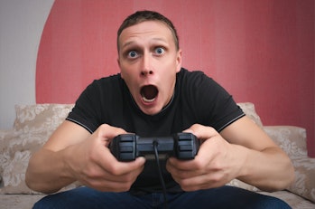 The surprised and enthusiastic gamer man with a joystick in his hand is sitting on a sofa with ...
