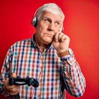 Senior handsome hoary gamer man playing video game using joystick and headphones serious face thinki...