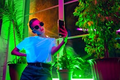 A woman with sunglasses on takes a selfie inside a neon-lit, plant-filled room. The January 2022 new...