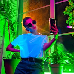 A woman with sunglasses on takes a selfie inside a neon-lit, plant-filled room. The January 2022 new...