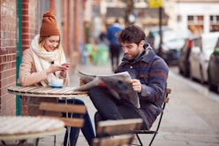 Couple On Date Sitting Outside Coffee Shop On High Street Using Mobile Phone And Reading Newspaper