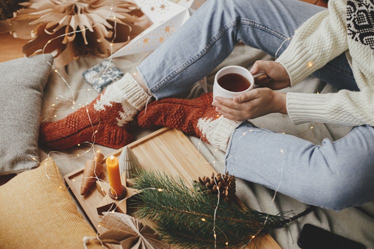 Woman hands with warm cup of tea relaxing on soft bed with festive decorations and pillows in scandi...