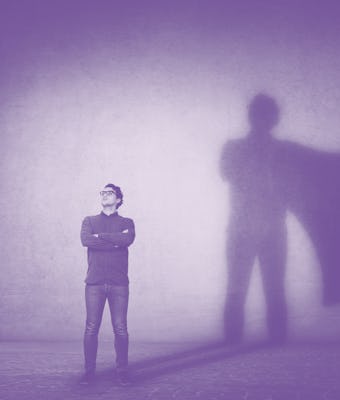 Brave man keeps arms crossed, looks confident, casting a superhero with cape shadow on the wall. Amb...