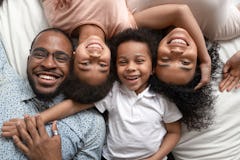 Happy mixed race family of four lying on bed cuddling, smiling african american dad mom and little k...