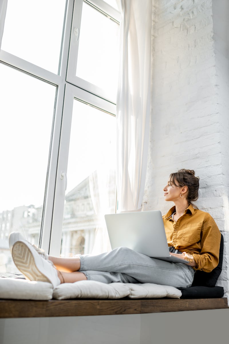 Young woman dressed casually working on laptop while sitting on the window sill at home. Work from h...