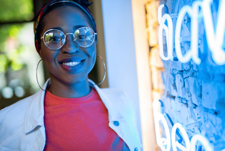 Happy young african american lady standing inside cafe near wall with neon light